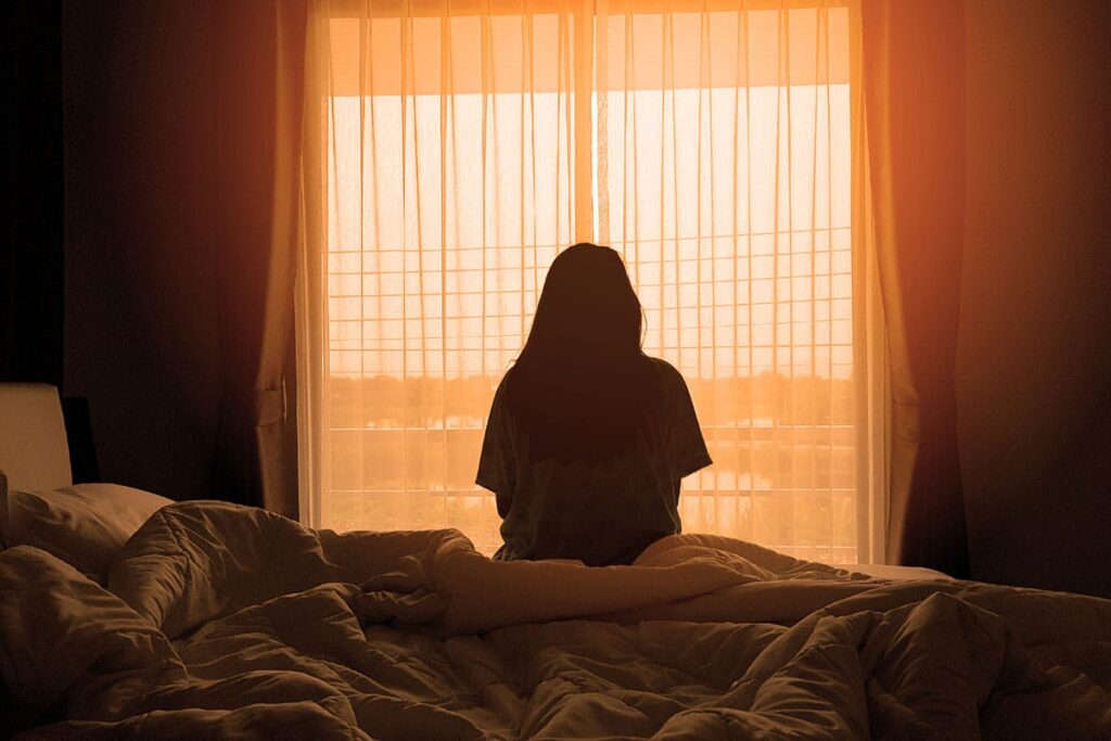 a silhouette of a person sitting on a bed framed by a large window streaming light while wondering what causes bipolar disorder